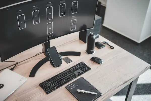 All black computer monitor, wireless keyboard, speaker, water bottle, wallet, ID, wireless mouse, phone, notepad on a wooden table. A white pen is on the notepad and part of a MacBook is visible to the left part of the picture.