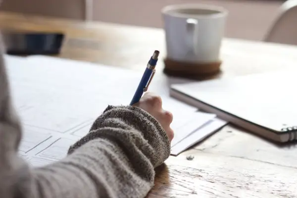 A person's had holding a pen and writing on paper with a cup in the background