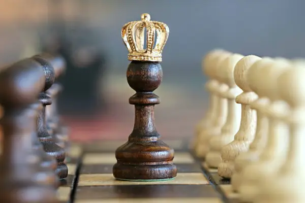 A brown wooden pawn chess piece with a small golden crown on top of it with brown pawns lined to the left and white pawns lined to the right