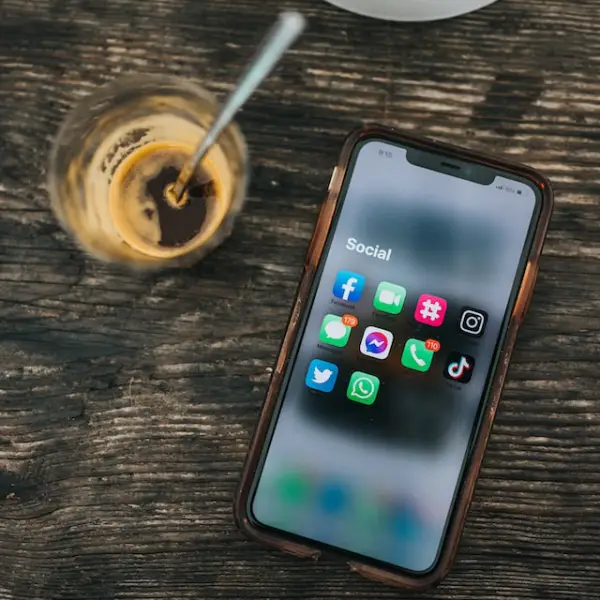 An iPhone displaying social media icons on the screen and a nearly empty glass of coffee with a spoon beside the phone
