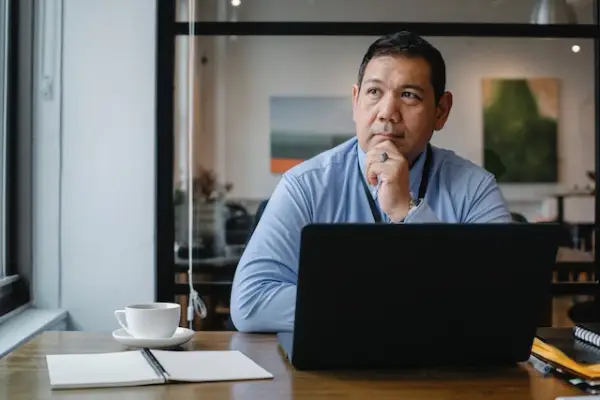 A man in a workplace in a thinking pose with his left hand below his chin and looking to his right. A laptop, cup, and notepad sit on a table he is leaning on.