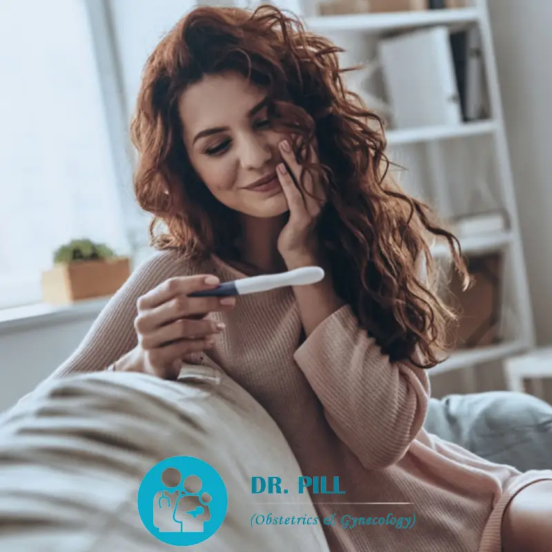 A smiling woman looking at a pregnancy test in her right hand with her left hand on her left cheek