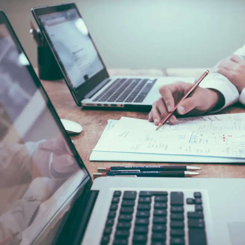 A person's hand holding a pencil writing on paper with two laptops on either side symbolizing person giving web development advice to client