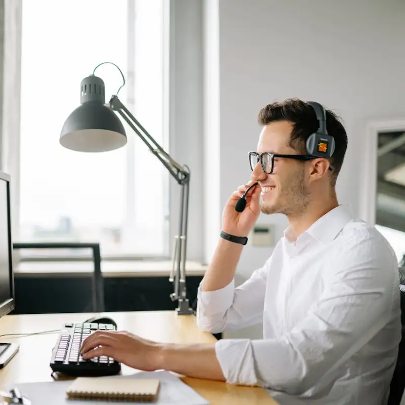 A smiling man with glasses on headphones on with his right hand on the headphone microphone and left hand on a computer keyboard on a desk providing customer support